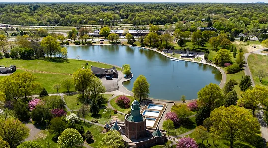 Aerial view of a park with a pond, green trees, flowering shrubs, and a historic building.