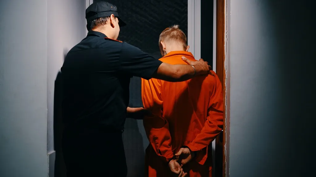 Police officer escorting a handcuffed individual in an orange jumpsuit through a doorway.