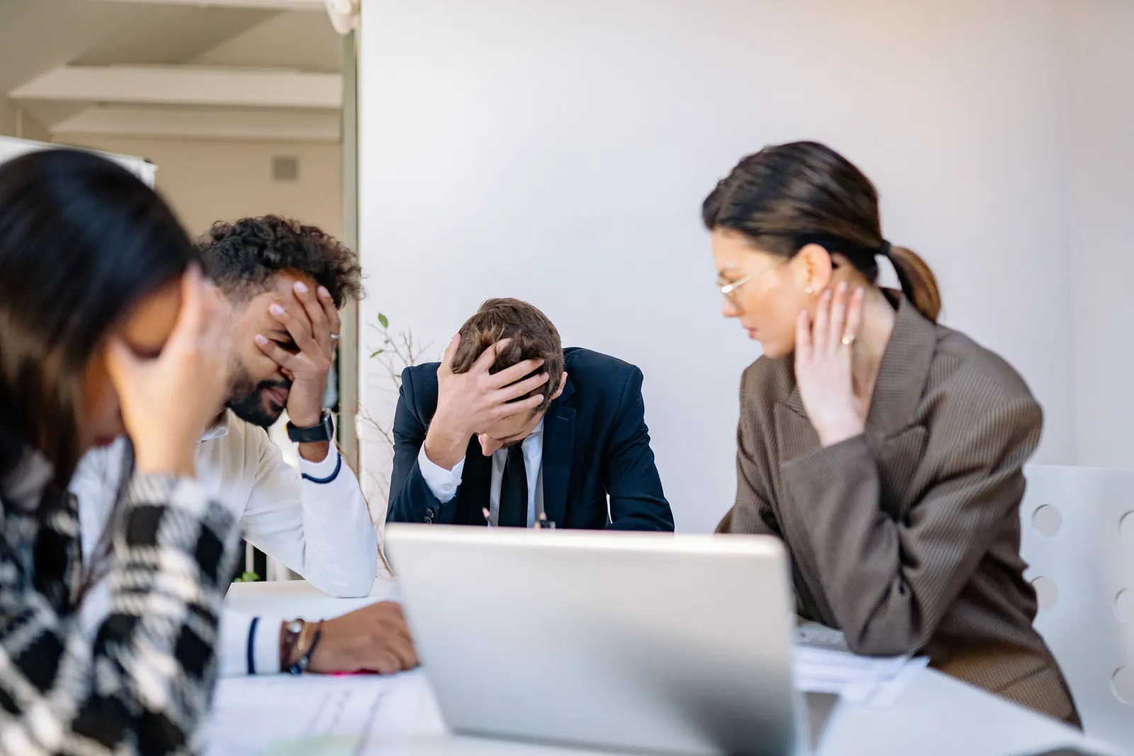 Four diverse business professionals seated at a table, visibly stressed with heads in hands.